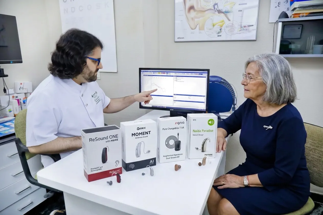 Un audioprotesista muestra los resultados en pantalla a una paciente, con varios modelos de audífono expuestos sobre la mesa