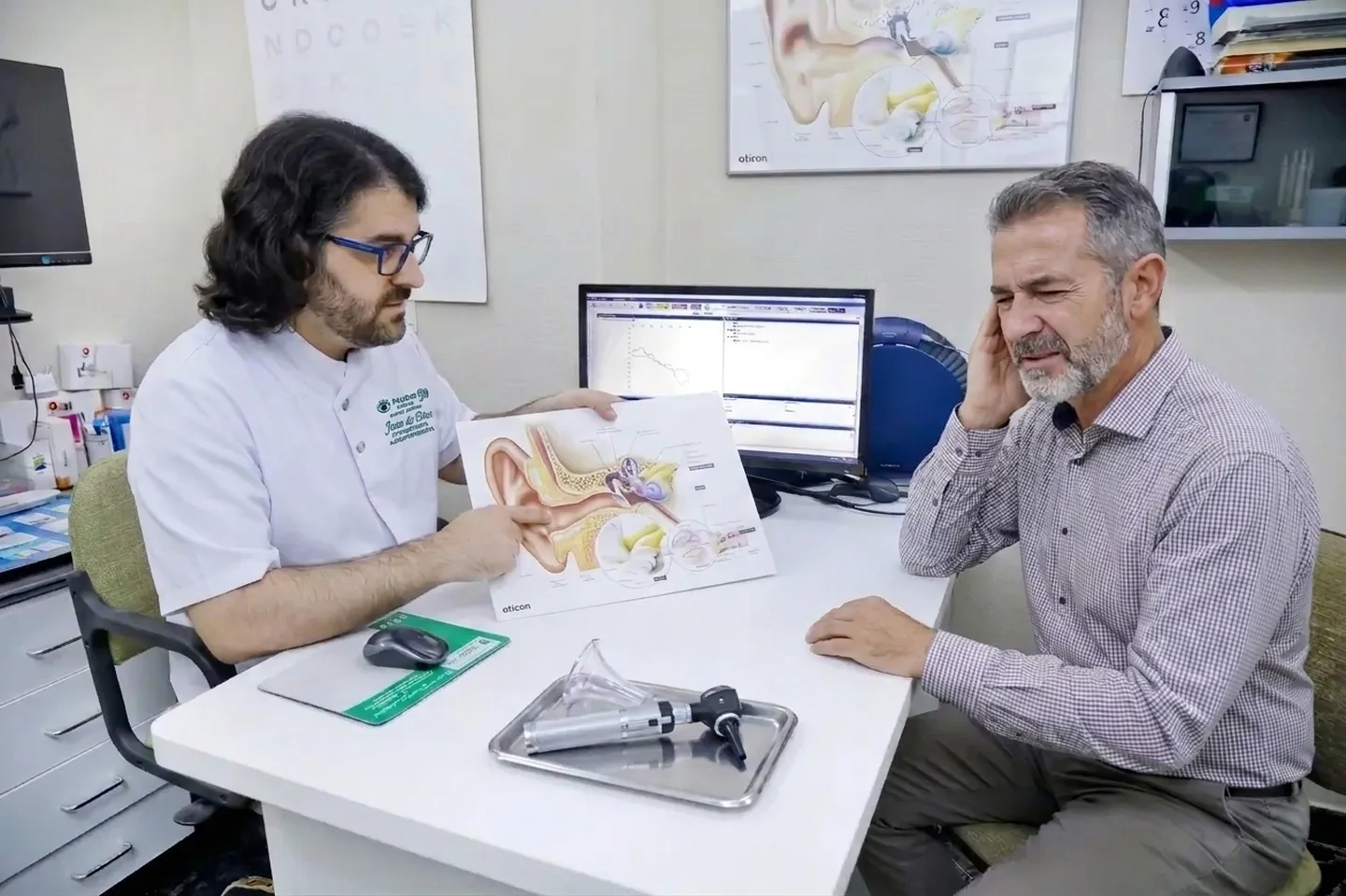 Un audioprotesista explica con una lámina anatómica del oído a un paciente que se toca la oreja